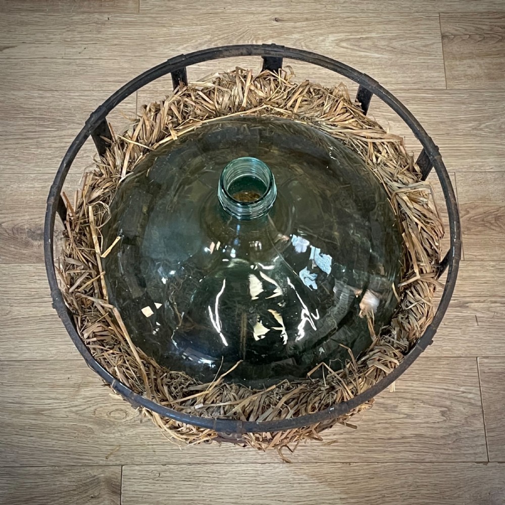 A large green glass demijohn bottle resting inside a natural straw-lined iron basket frame. View V