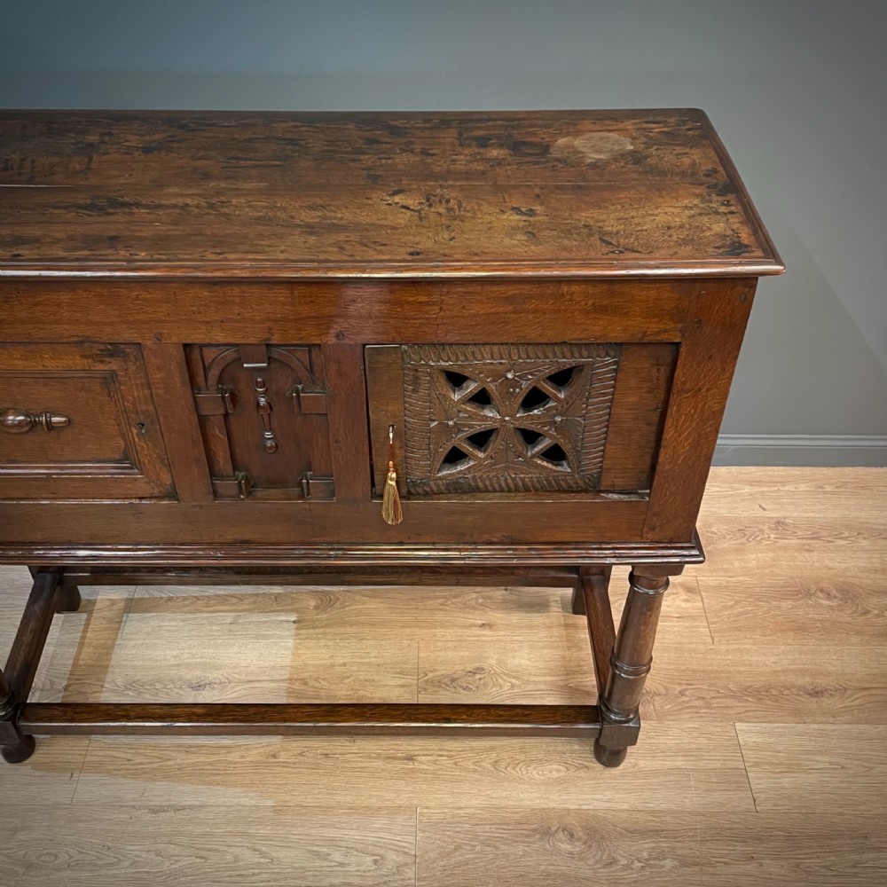 An antique dark oak livery cupboard featuring a carved pierced ventilation panel, central arch motif, turned legs, and a lower stretcher base. View V