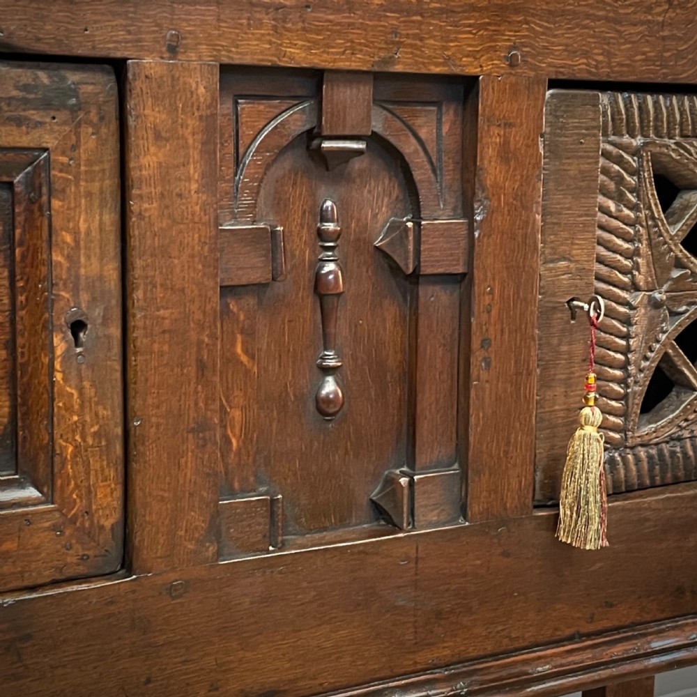 An antique dark oak livery cupboard featuring a carved pierced ventilation panel, central arch motif, turned legs, and a lower stretcher base. View VI