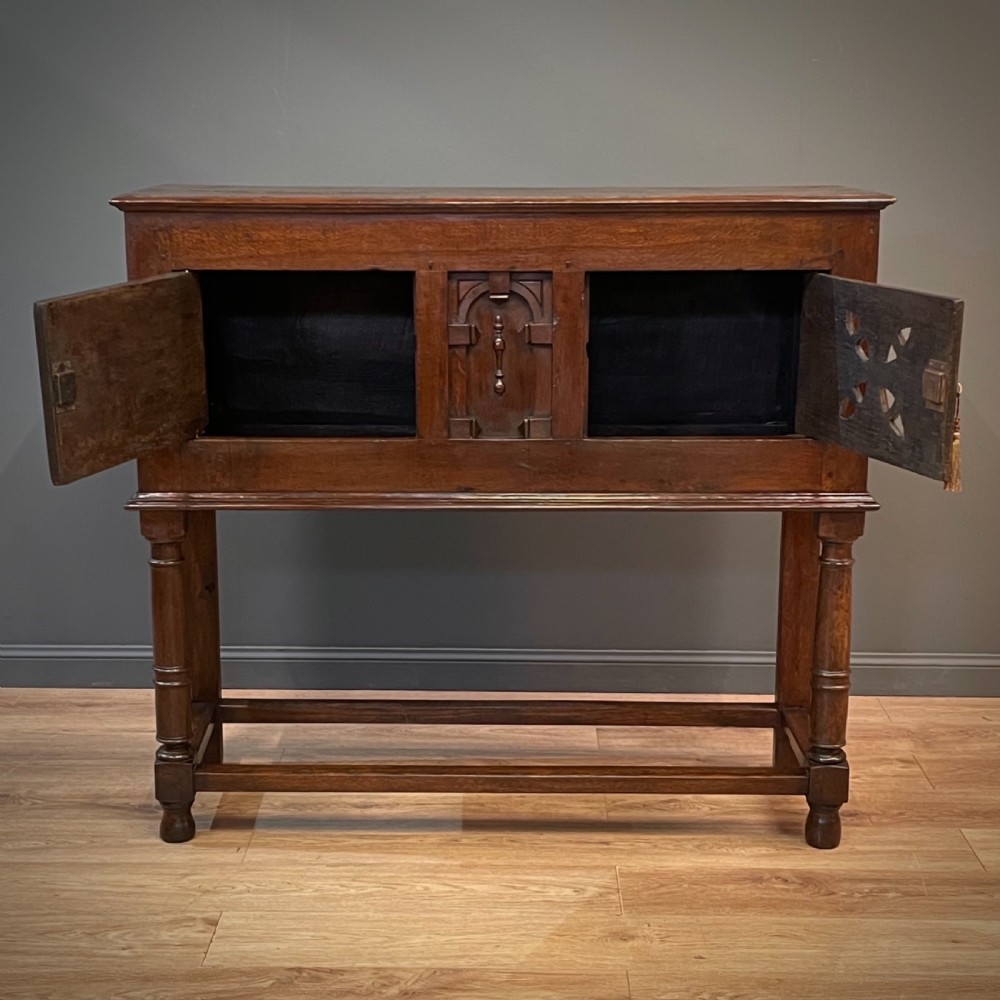 An antique dark oak livery cupboard featuring a carved pierced ventilation panel, central arch motif, turned legs, and a lower stretcher base. View XI