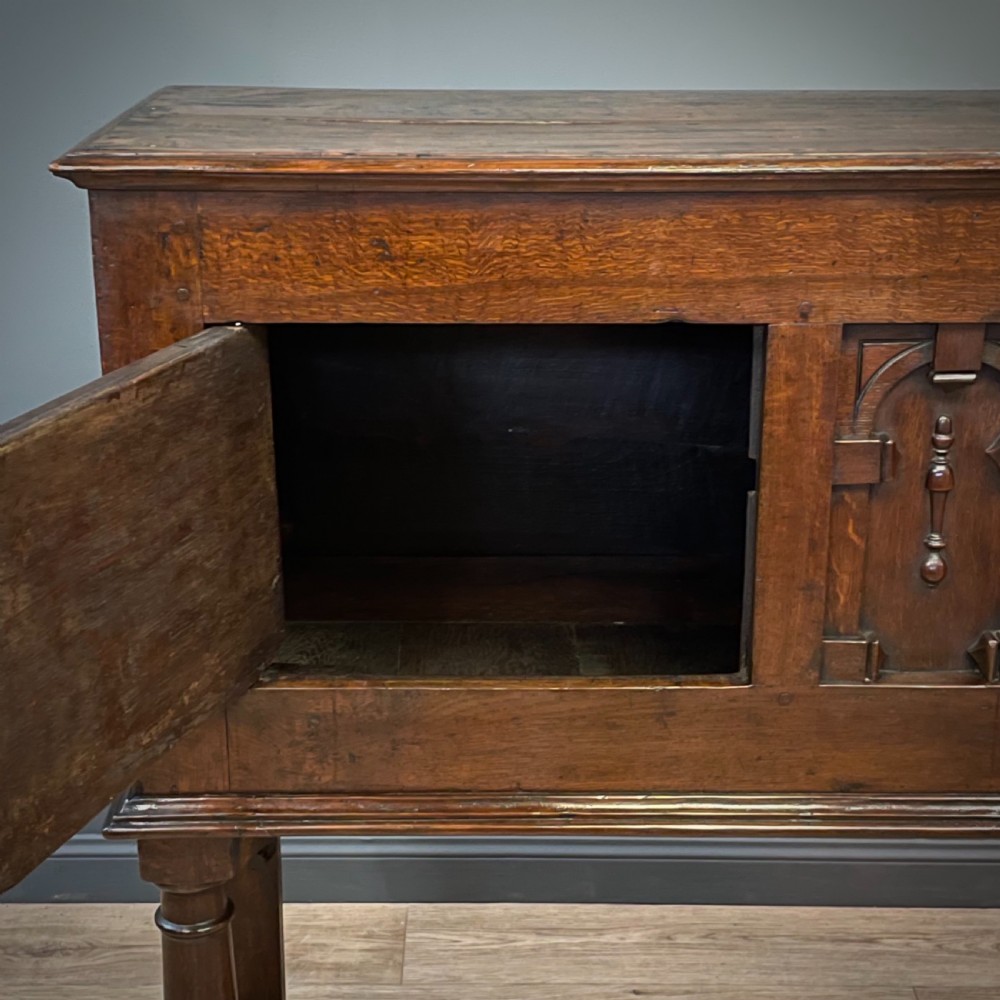 An antique dark oak livery cupboard featuring a carved pierced ventilation panel, central arch motif, turned legs, and a lower stretcher base. View XII
