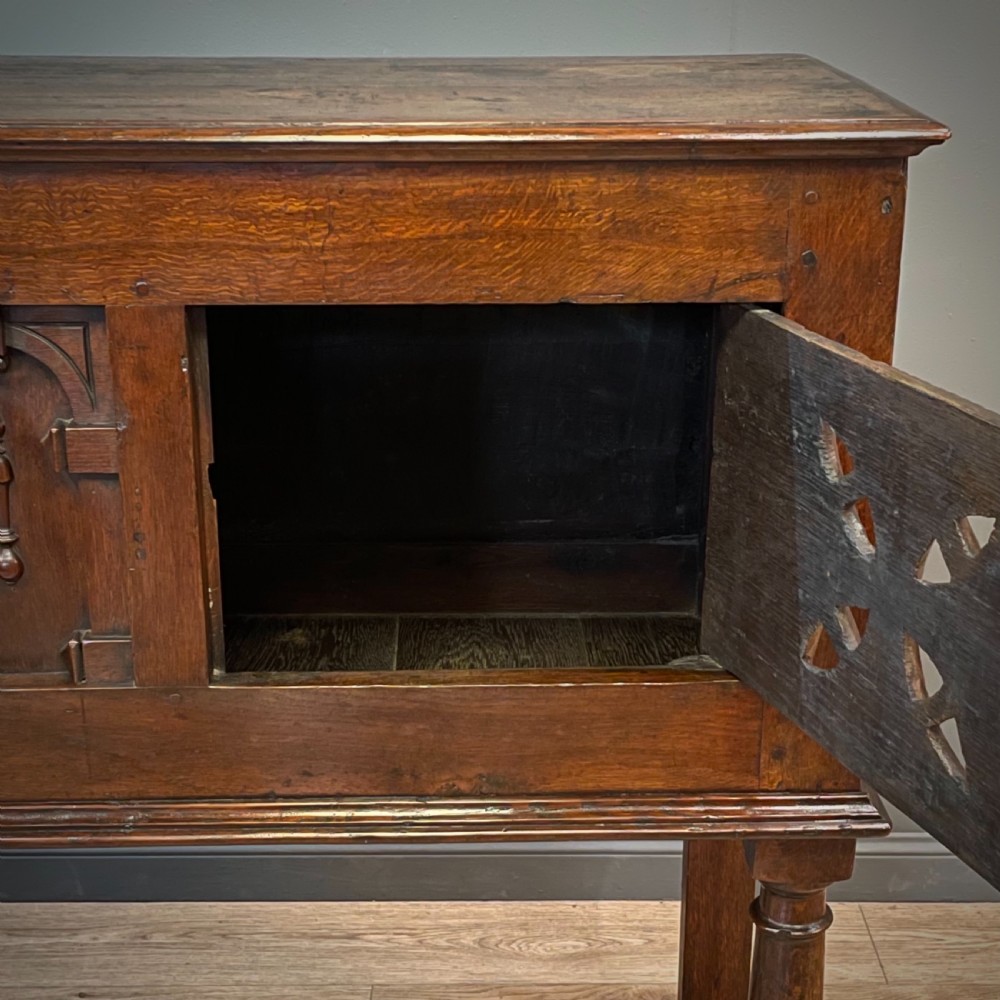 An antique dark oak livery cupboard featuring a carved pierced ventilation panel, central arch motif, turned legs, and a lower stretcher base. View XIII