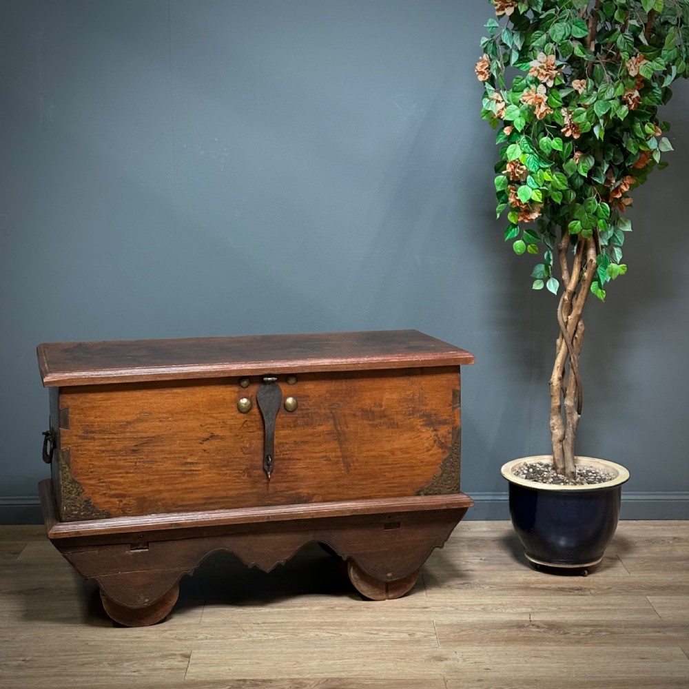 A rustic warm brown teak marriage chest featuring iron latch hardware, brass corner brackets, and a raised wooden base supported by four solid wooden wheels.