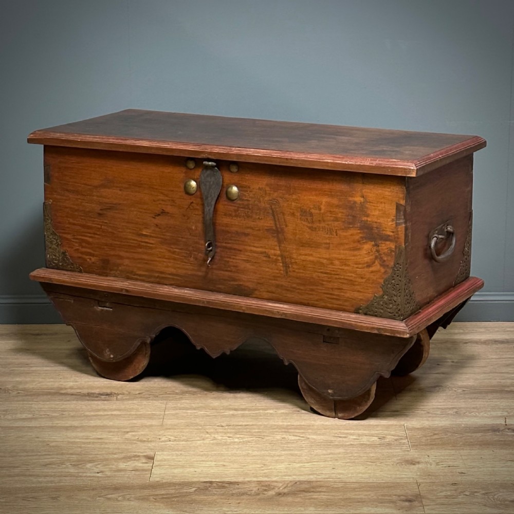 A rustic warm brown teak marriage chest featuring iron latch hardware, brass corner brackets, and a raised wooden base supported by four solid wooden wheels. View III