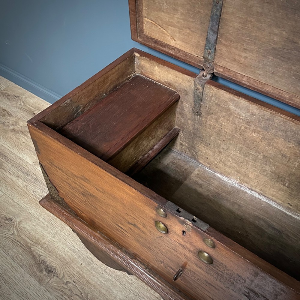 A rustic warm brown teak marriage chest featuring iron latch hardware, brass corner brackets, and a raised wooden base supported by four solid wooden wheels. View V