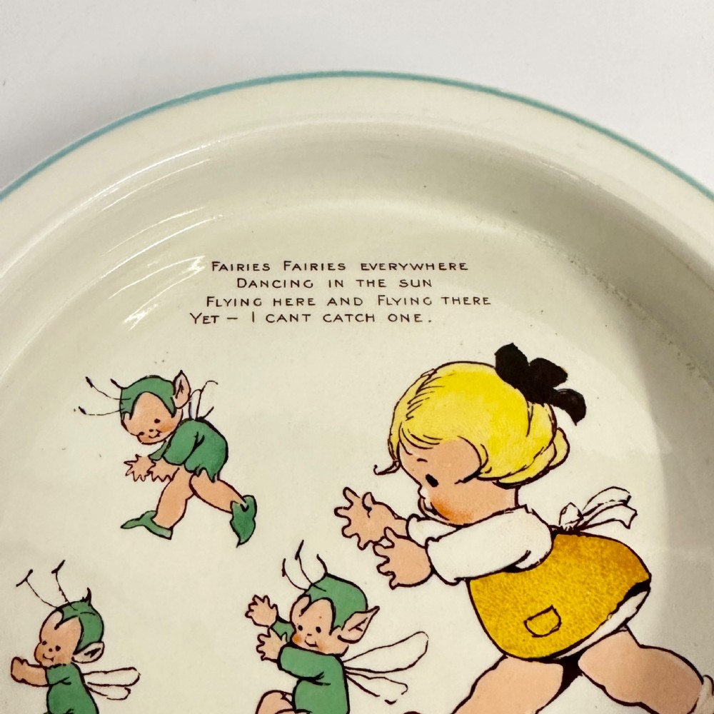 A circular cream glazed ceramic deep baby plate with green rim trim and a central illustration of a child and fairies by Mabel Lucie Attwell. View II
