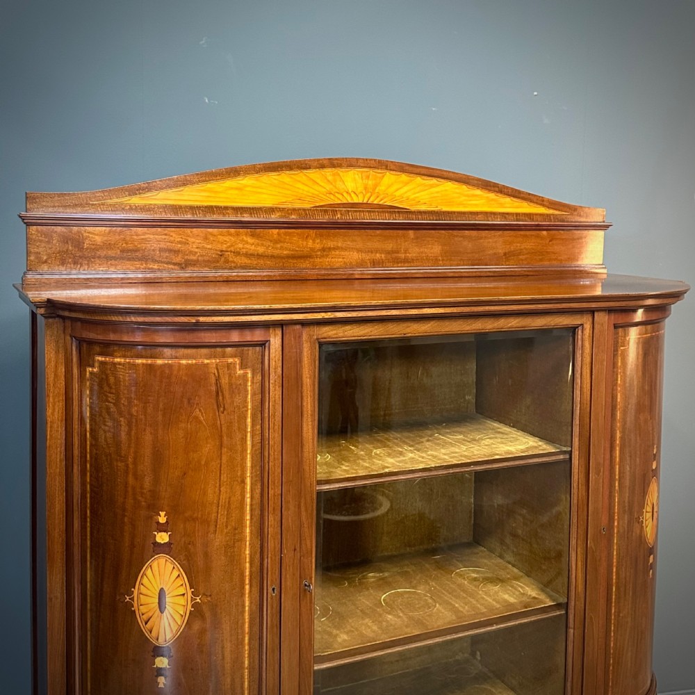 An Edwardian bow-fronted mahogany display cabinet featuring a central glazed door flanked by satinwood oval patera marquetry panels and an arched pediment. View II
