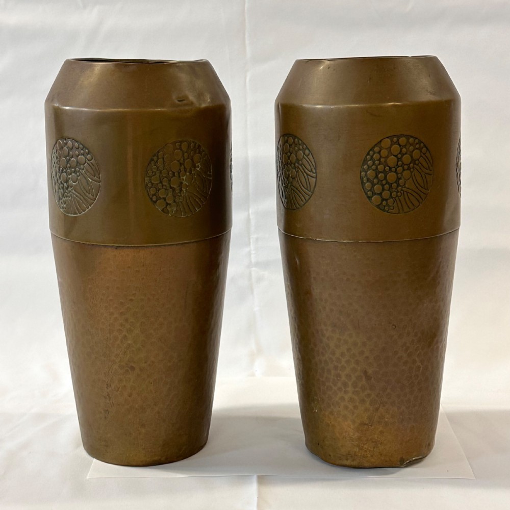 A pair of cylindrical brass vases featuring hand-hammered lower bodies and circular embossed floral medallions around the upper rim. View V