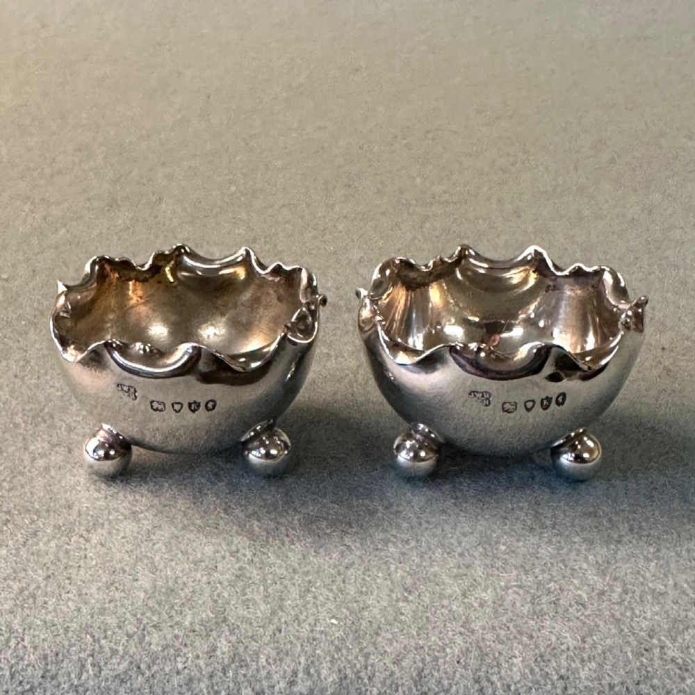 A pair of Victorian sterling silver open salt cellars featuring crimped rims, stamped hallmarks, and raised on three ball feet.