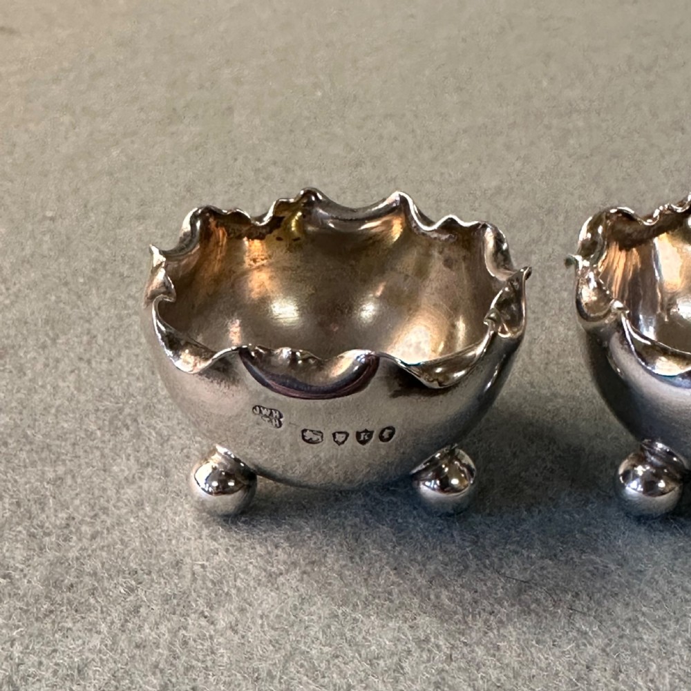 A pair of Victorian sterling silver open salt cellars featuring crimped rims, stamped hallmarks, and raised on three ball feet. View II