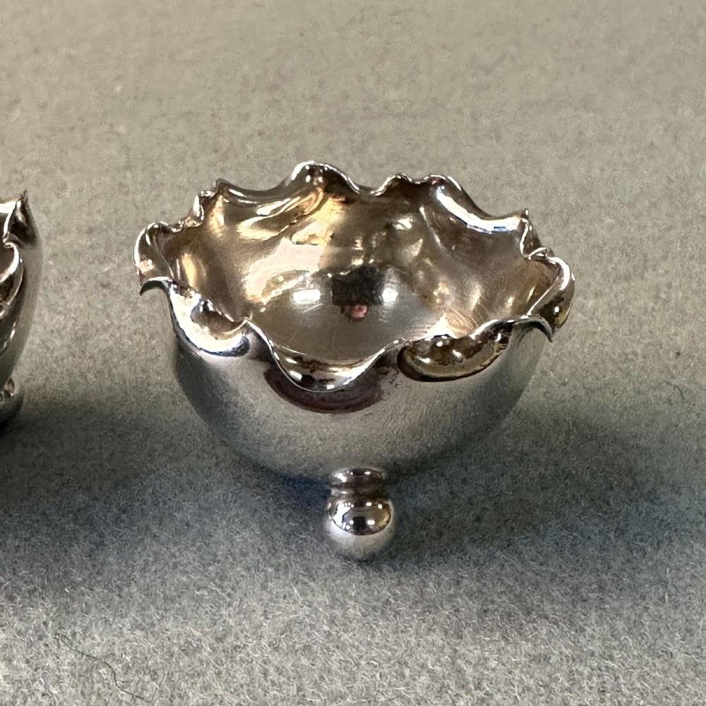 A pair of Victorian sterling silver open salt cellars featuring crimped rims, stamped hallmarks, and raised on three ball feet. View V