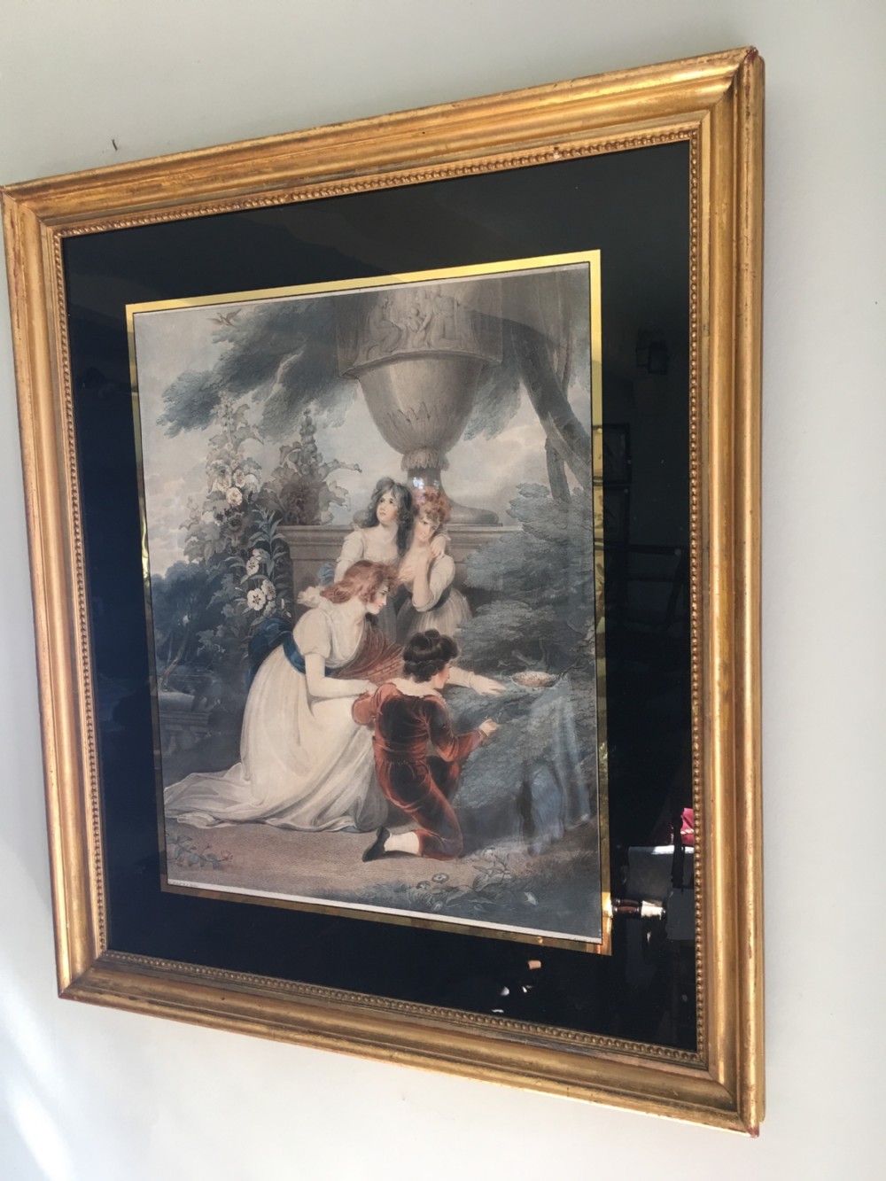 A framed Regency coloured mezzotint depicting figures discovering a bird nest in a garden, mounted within a black and gold glass border and a gilded frame.