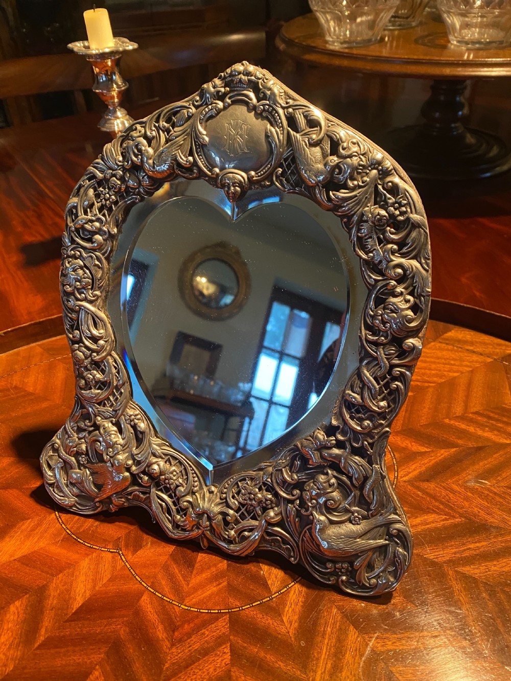 A heart-shaped silver easel mirror featuring intricate pierced scrollwork, floral motifs, bird details, and an engraved top cartouche.