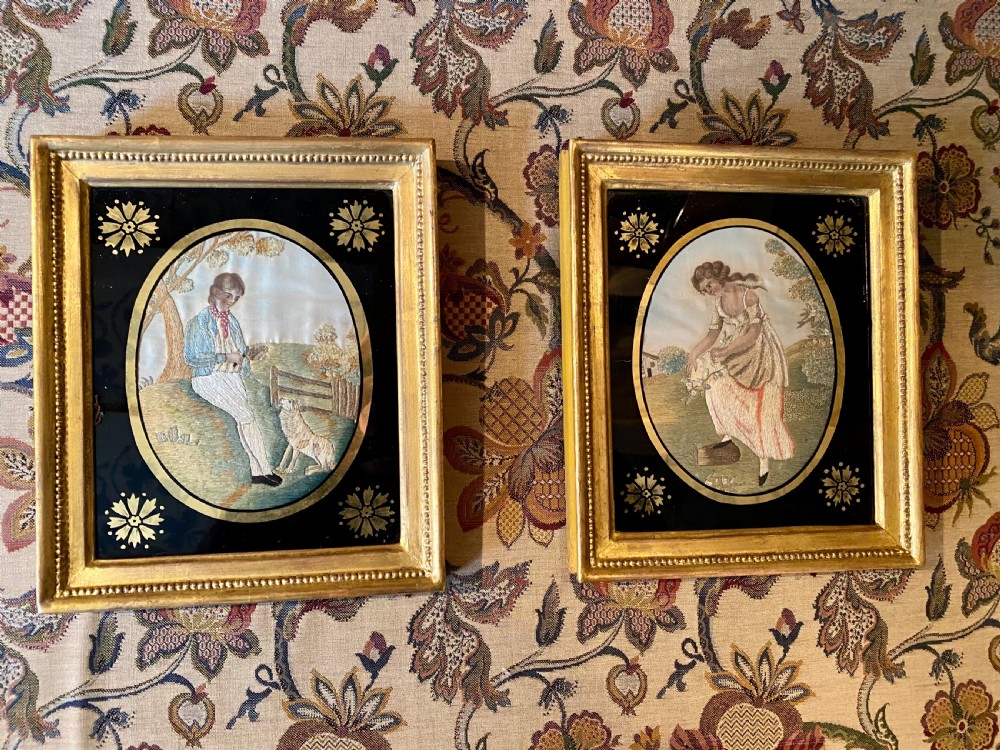 A pair of rectangular gold giltwood framed antique pictures featuring oval silk embroidery of pastoral figures against black glass borders.