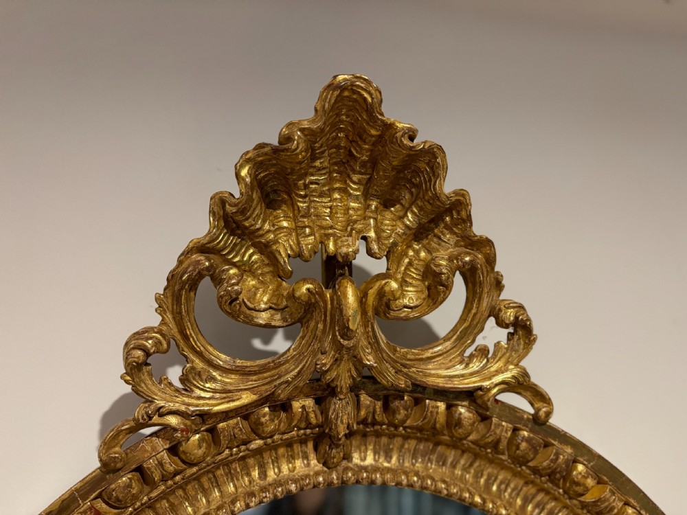 A George III period oval wall mirror featuring a carved giltwood frame with cresting detail and molded inner borders. View II
