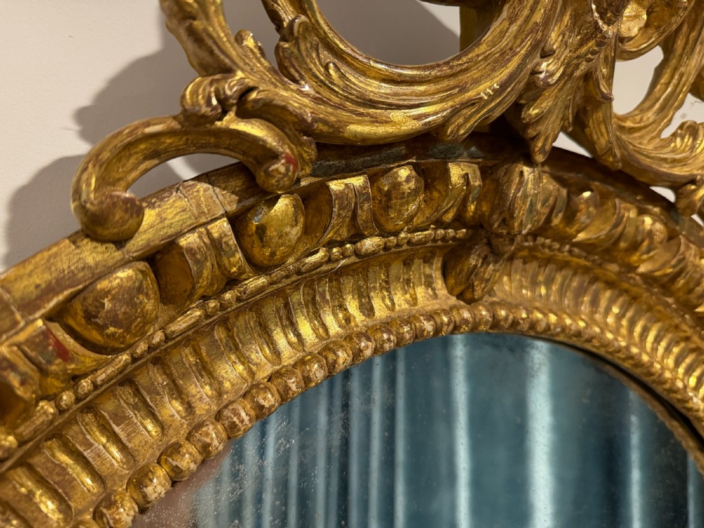 A George III period oval wall mirror featuring a carved giltwood frame with cresting detail and molded inner borders. View III