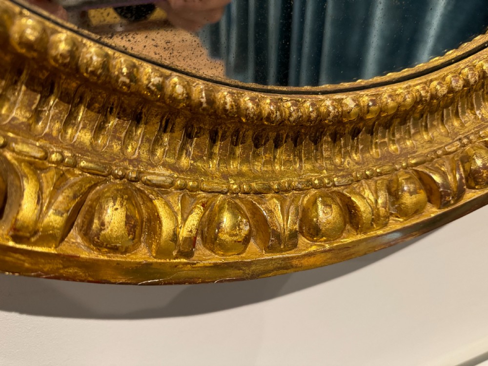 A George III period oval wall mirror featuring a carved giltwood frame with cresting detail and molded inner borders. View IV