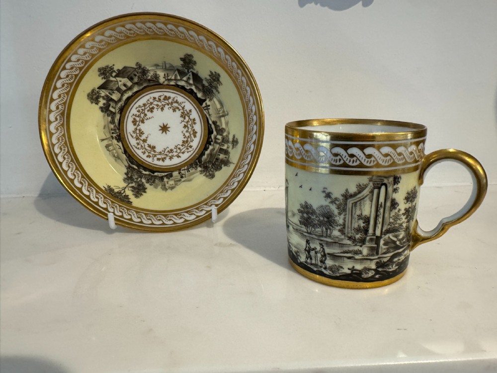 A straight-sided French porcelain coffee cup and saucer featuring hand-painted sepia landscape scenes, a pale yellow ground, and wide gold leaf borders.