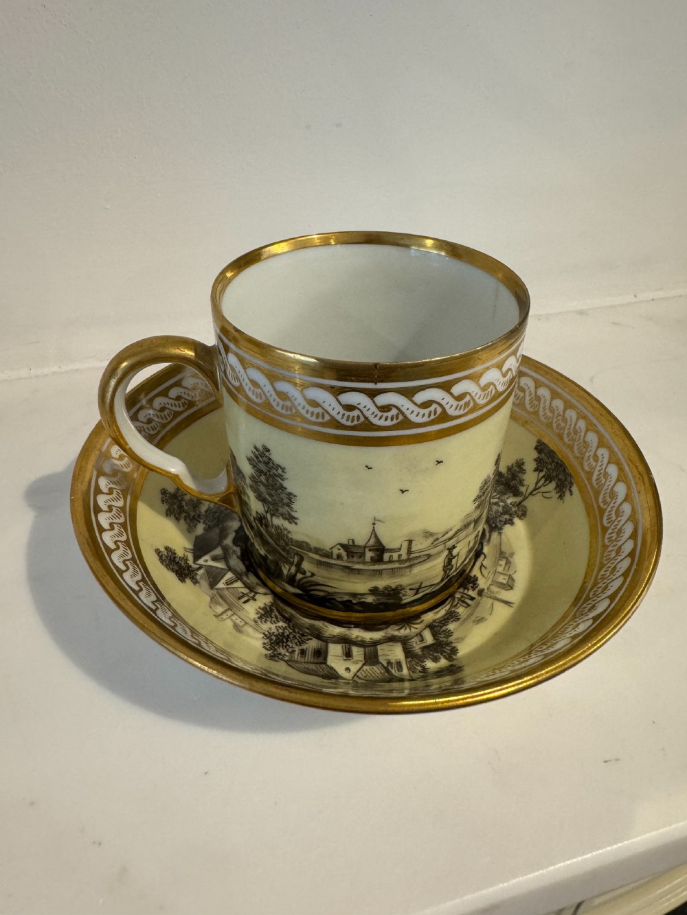 A straight-sided French porcelain coffee cup and saucer featuring hand-painted sepia landscape scenes, a pale yellow ground, and wide gold leaf borders. View II