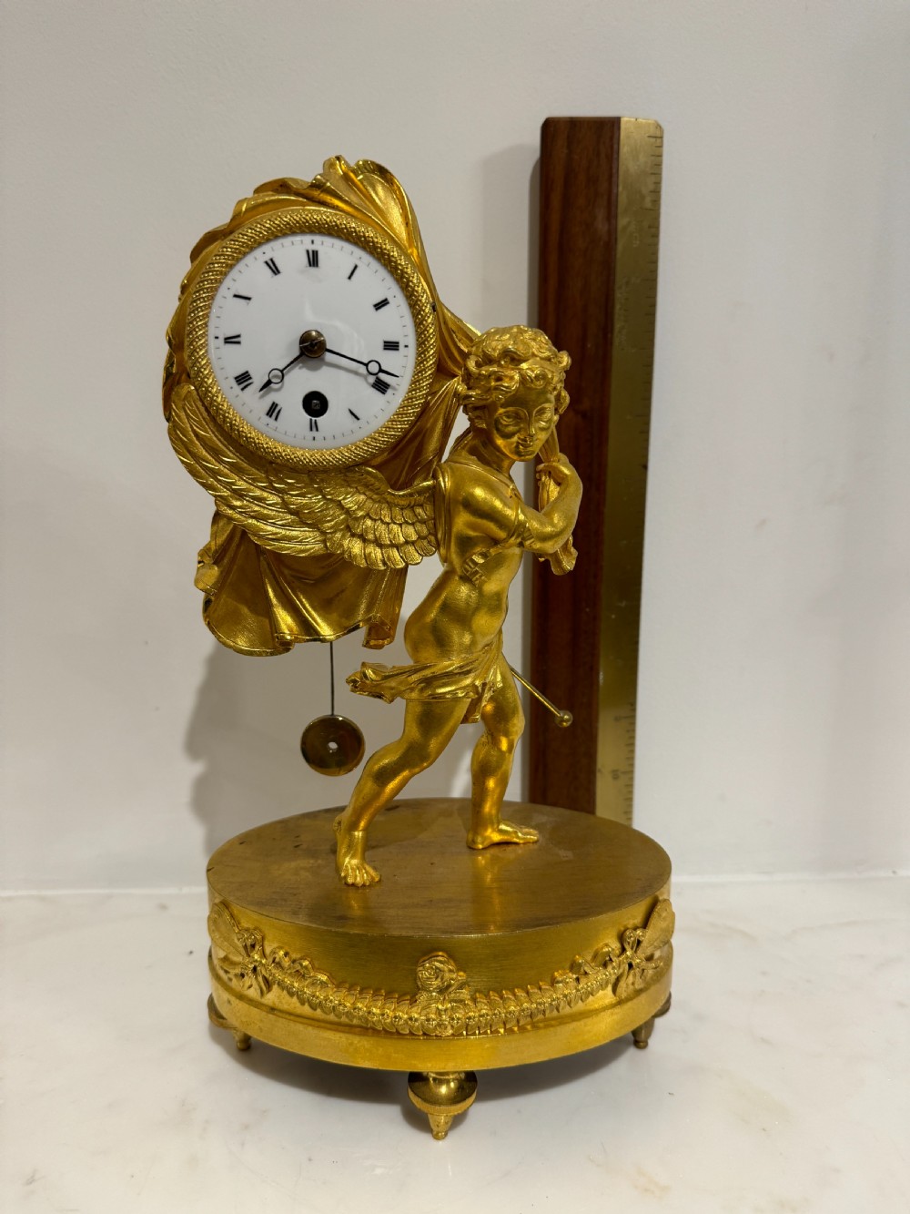 A French Empire gilt bronze table clock featuring a sculpted winged cherub figure supporting a circular white enamel dial set on an oval decorated base. View II