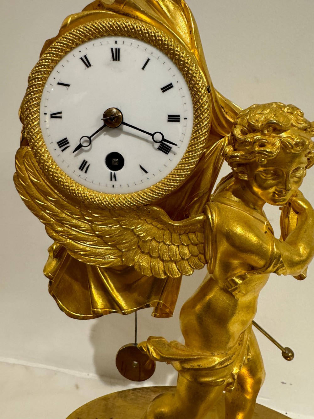 A French Empire gilt bronze table clock featuring a sculpted winged cherub figure supporting a circular white enamel dial set on an oval decorated base. View III