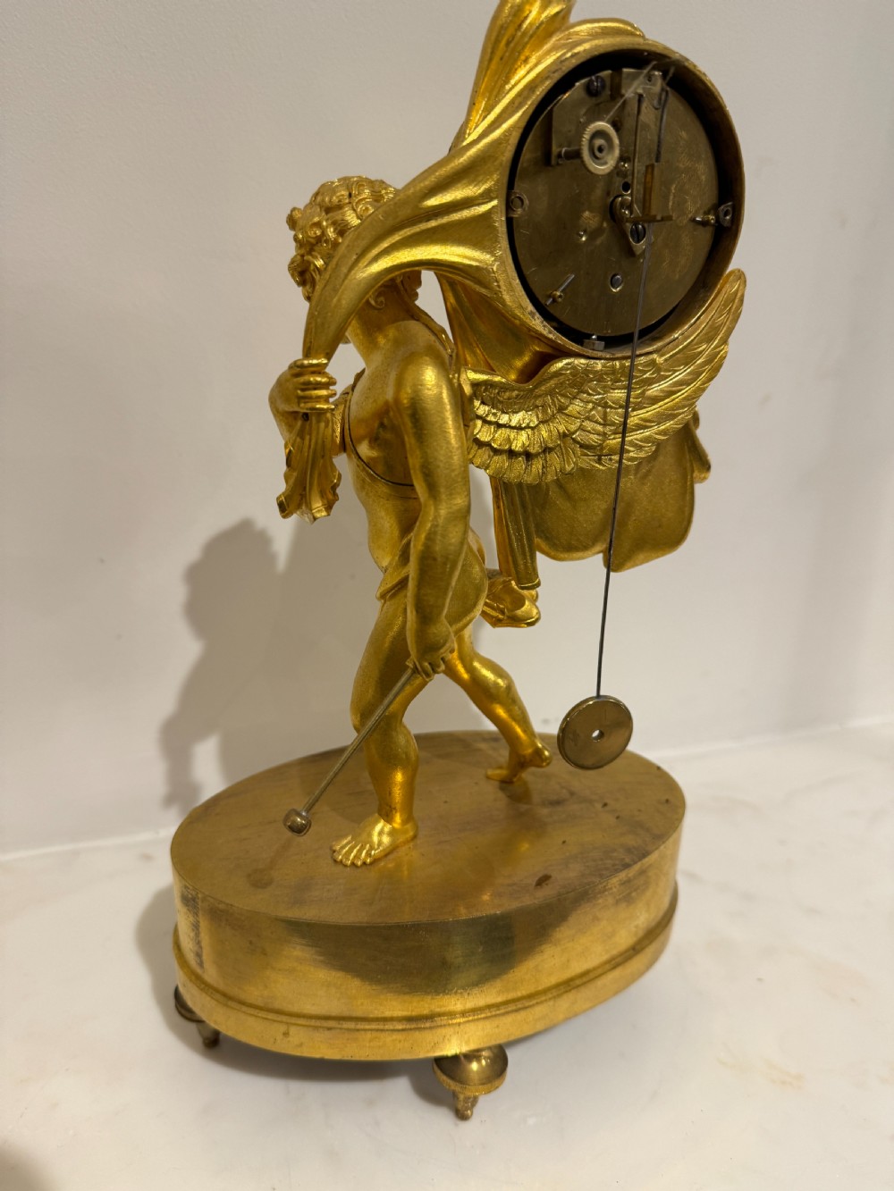 A French Empire gilt bronze table clock featuring a sculpted winged cherub figure supporting a circular white enamel dial set on an oval decorated base. View V