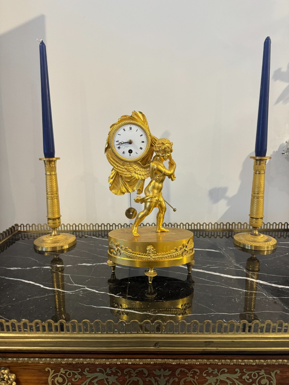 A French Empire gilt bronze table clock featuring a sculpted winged cherub figure supporting a circular white enamel dial set on an oval decorated base. View VII