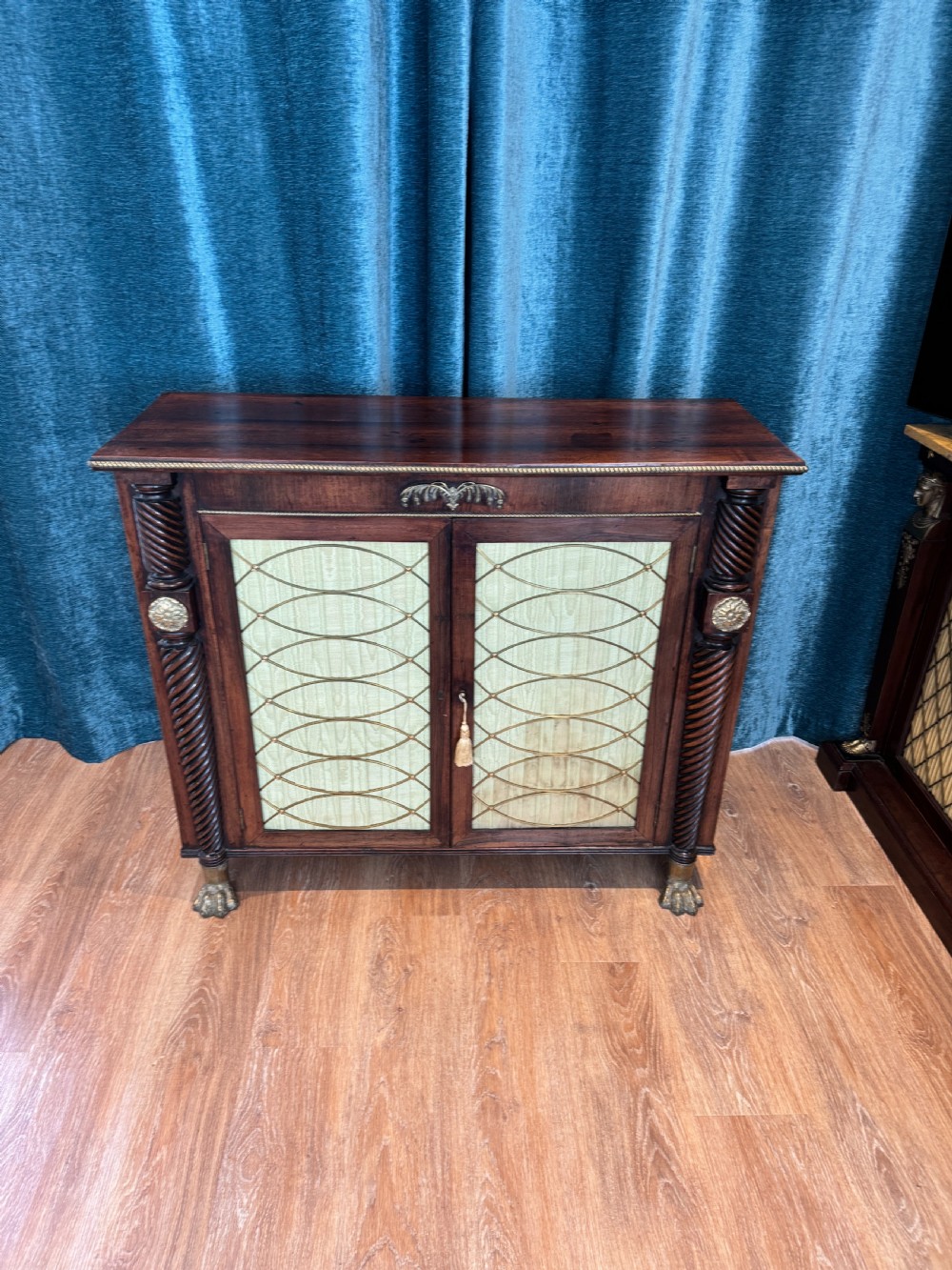 A Regency dark wood side cabinet featuring rope-twist side columns, brass paw feet, and wire-grille doors backed with pleated pale green fabric.