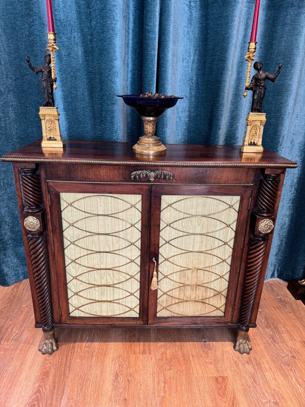A Regency dark wood side cabinet featuring rope-twist side columns, brass paw feet, and wire-grille doors backed with pleated pale green fabric. View II