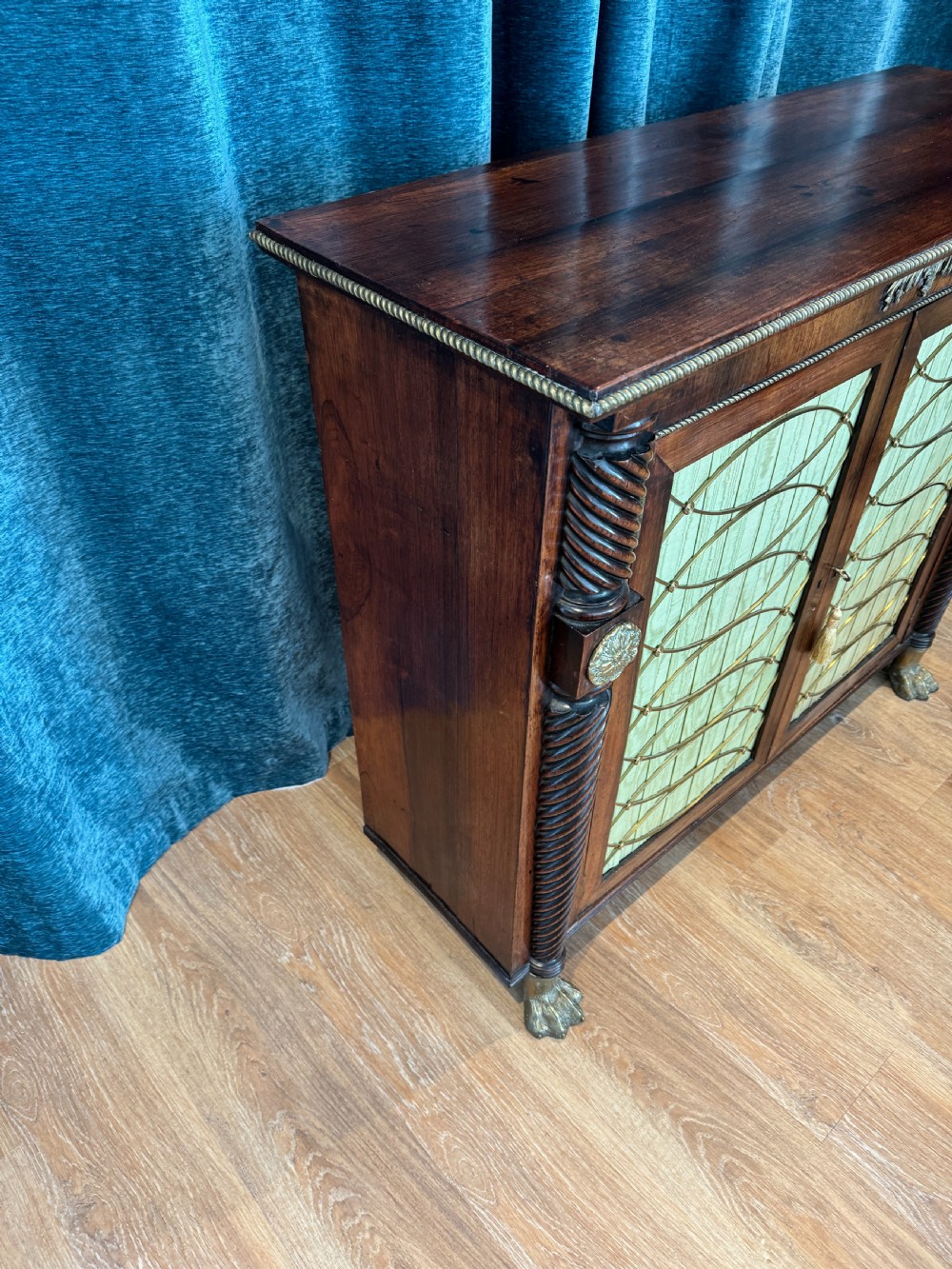 A Regency dark wood side cabinet featuring rope-twist side columns, brass paw feet, and wire-grille doors backed with pleated pale green fabric. View III