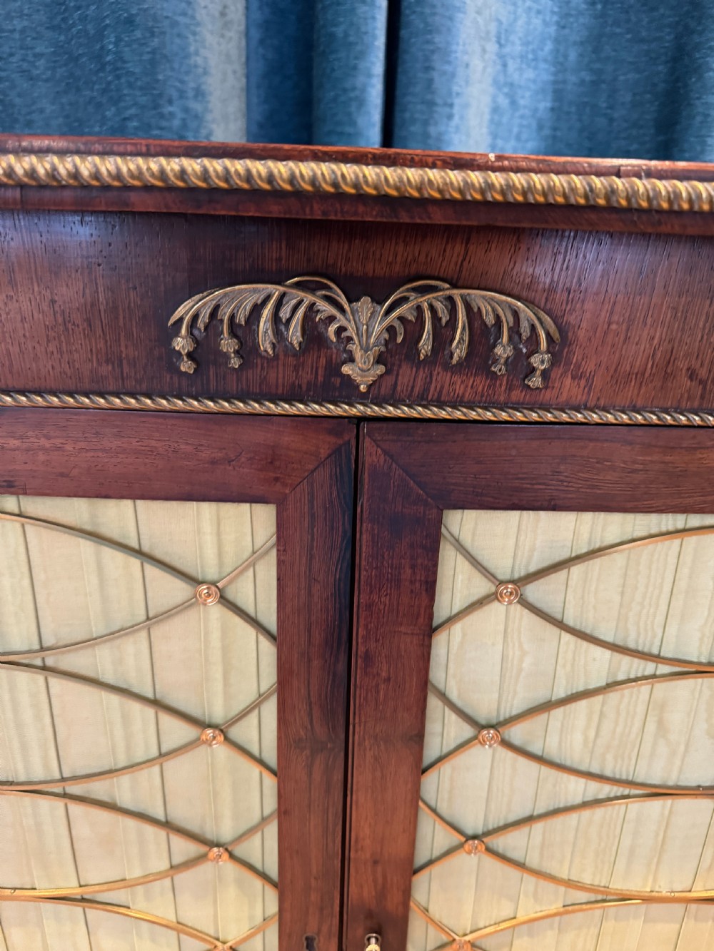 A Regency dark wood side cabinet featuring rope-twist side columns, brass paw feet, and wire-grille doors backed with pleated pale green fabric. View IV