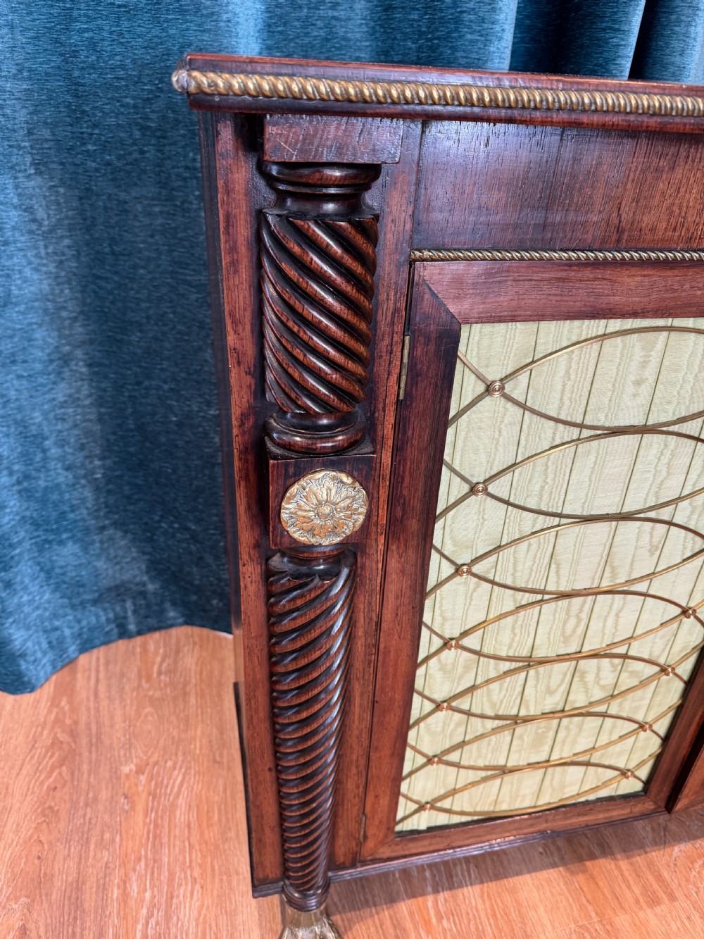 A Regency dark wood side cabinet featuring rope-twist side columns, brass paw feet, and wire-grille doors backed with pleated pale green fabric. View V