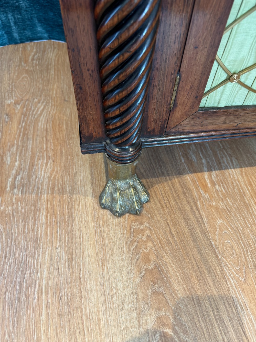 A Regency dark wood side cabinet featuring rope-twist side columns, brass paw feet, and wire-grille doors backed with pleated pale green fabric. View VI