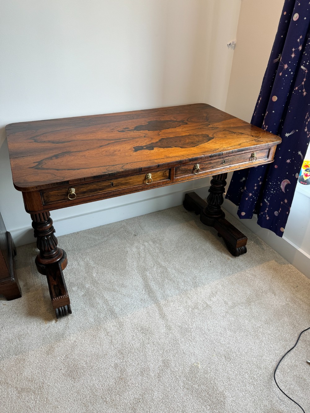 A Regency rosewood writing table featuring a highly figured top, two frieze drawers with brass lion-mask handles, and carved turned pedestal legs on trestle feet. View IV