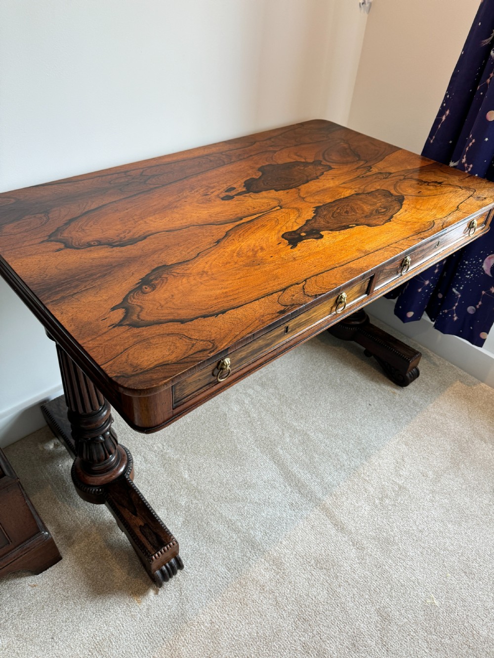 A Regency rosewood writing table featuring a highly figured top, two frieze drawers with brass lion-mask handles, and carved turned pedestal legs on trestle feet. View VII