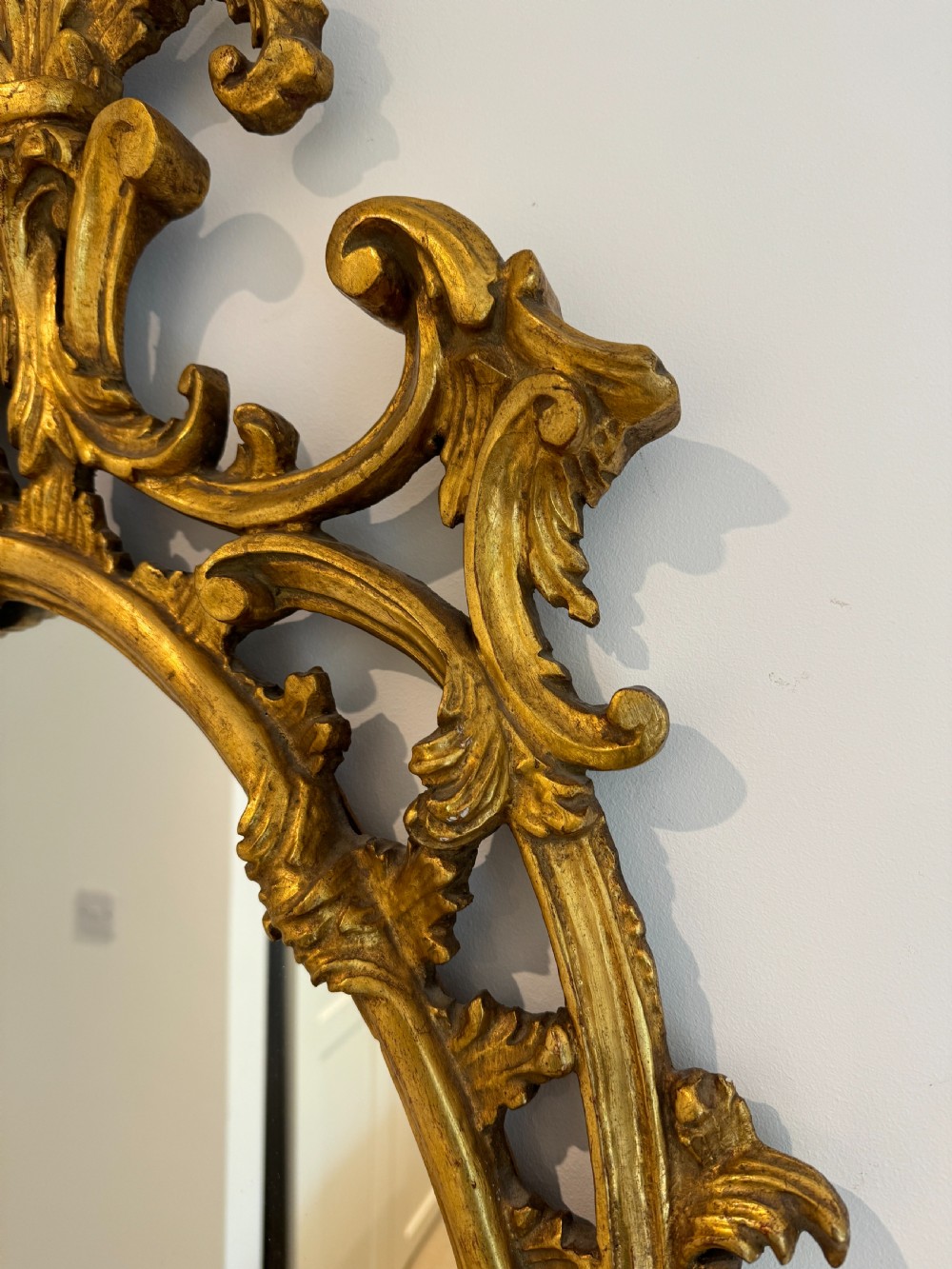 An ornate George III revival oval wall mirror featuring detailed carved giltwood scrollwork and an acanthus cresting. View II