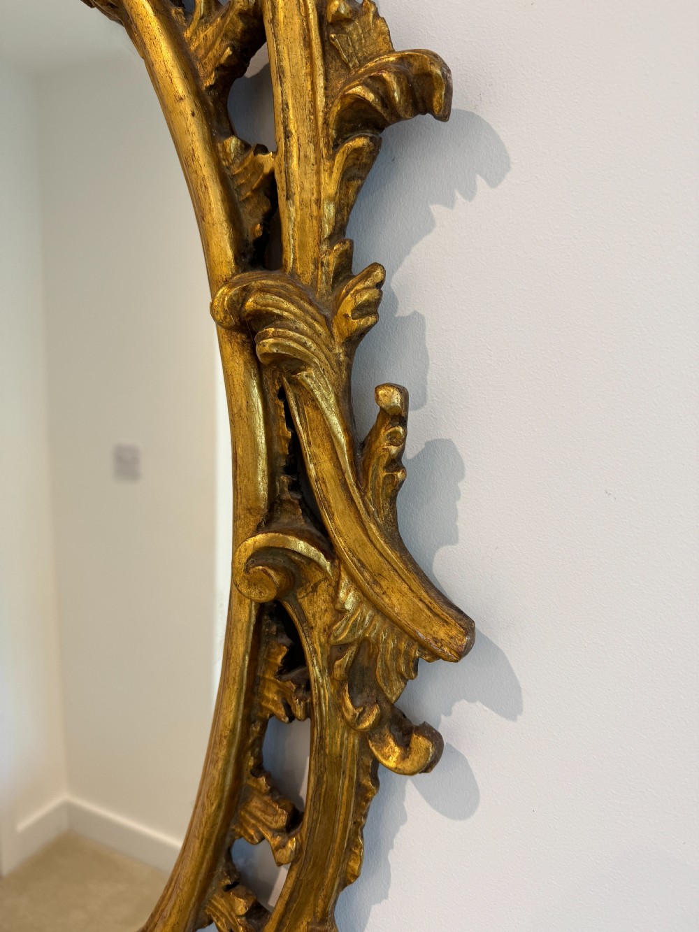 An ornate George III revival oval wall mirror featuring detailed carved giltwood scrollwork and an acanthus cresting. View IV