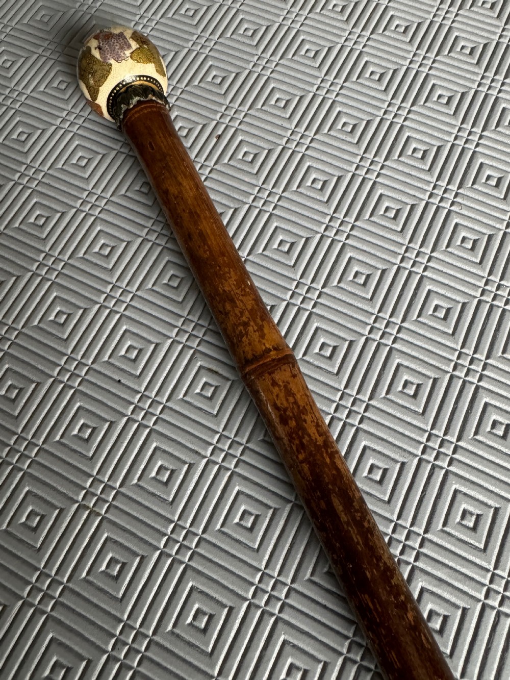 A dark brown bamboo walking stick featuring a cream and gold painted ceramic pommel handle. View III