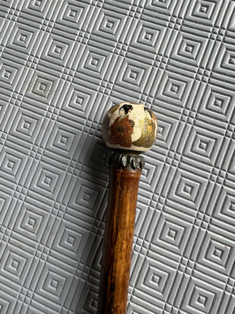 A dark brown bamboo walking stick featuring a cream and gold painted ceramic pommel handle. View V