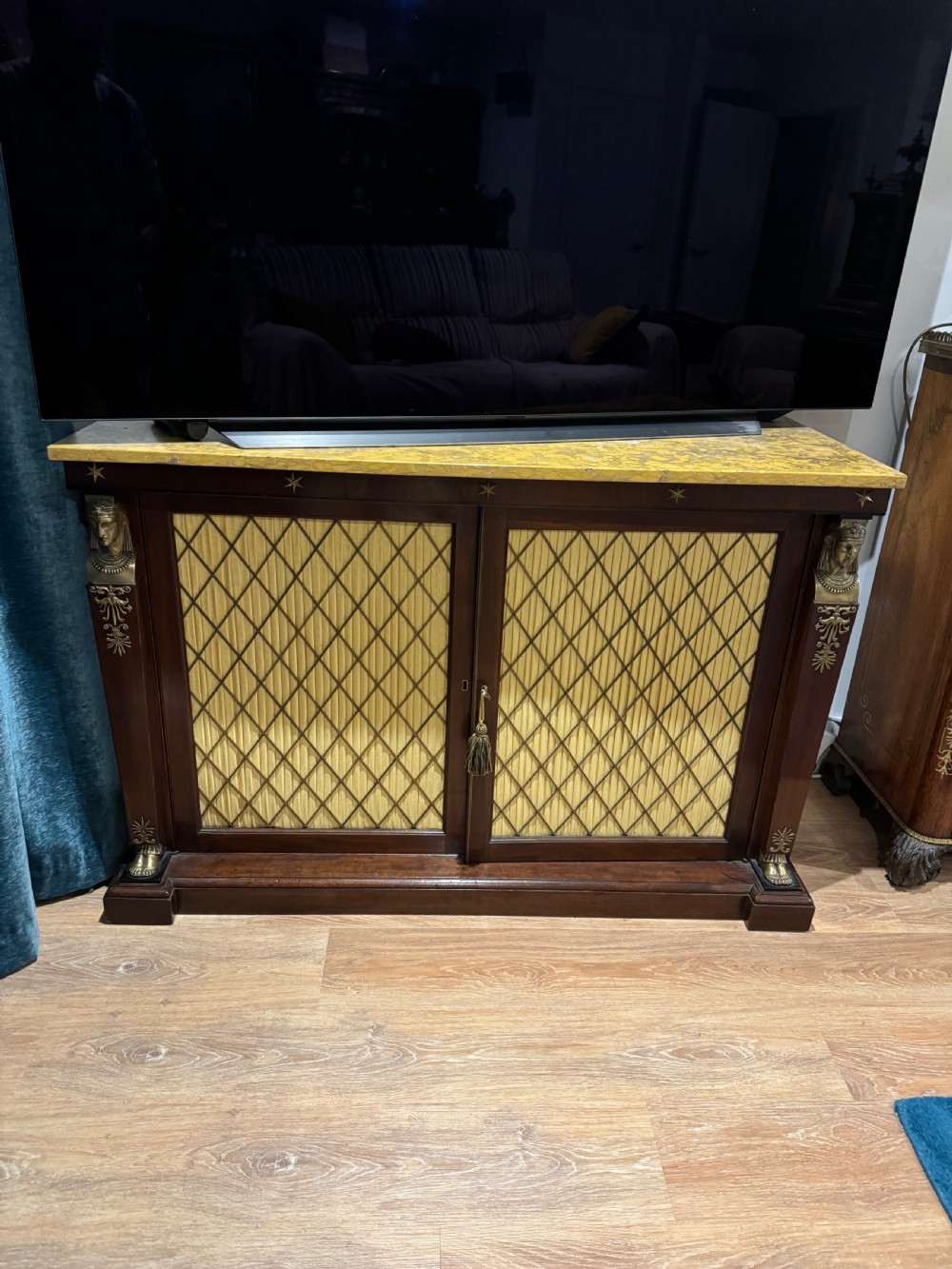 A Regency period dark mahogany side cabinet featuring an original yellow breche marble top, Egyptian revival gilt bronze mounts, and brass wire grille doors with pleated silk backing.