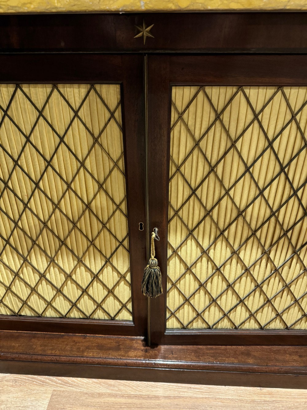 A Regency period dark mahogany side cabinet featuring an original yellow breche marble top, Egyptian revival gilt bronze mounts, and brass wire grille doors with pleated silk backing. View II