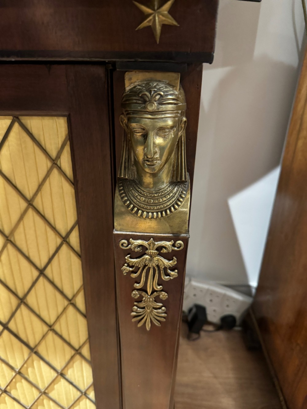 A Regency period dark mahogany side cabinet featuring an original yellow breche marble top, Egyptian revival gilt bronze mounts, and brass wire grille doors with pleated silk backing. View VII