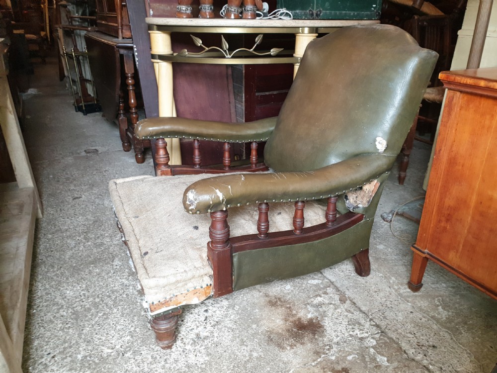 A deep Victorian Howard style armchair upholstered in worn olive green leather with turned wooden spindles under the armrests and turned front legs.