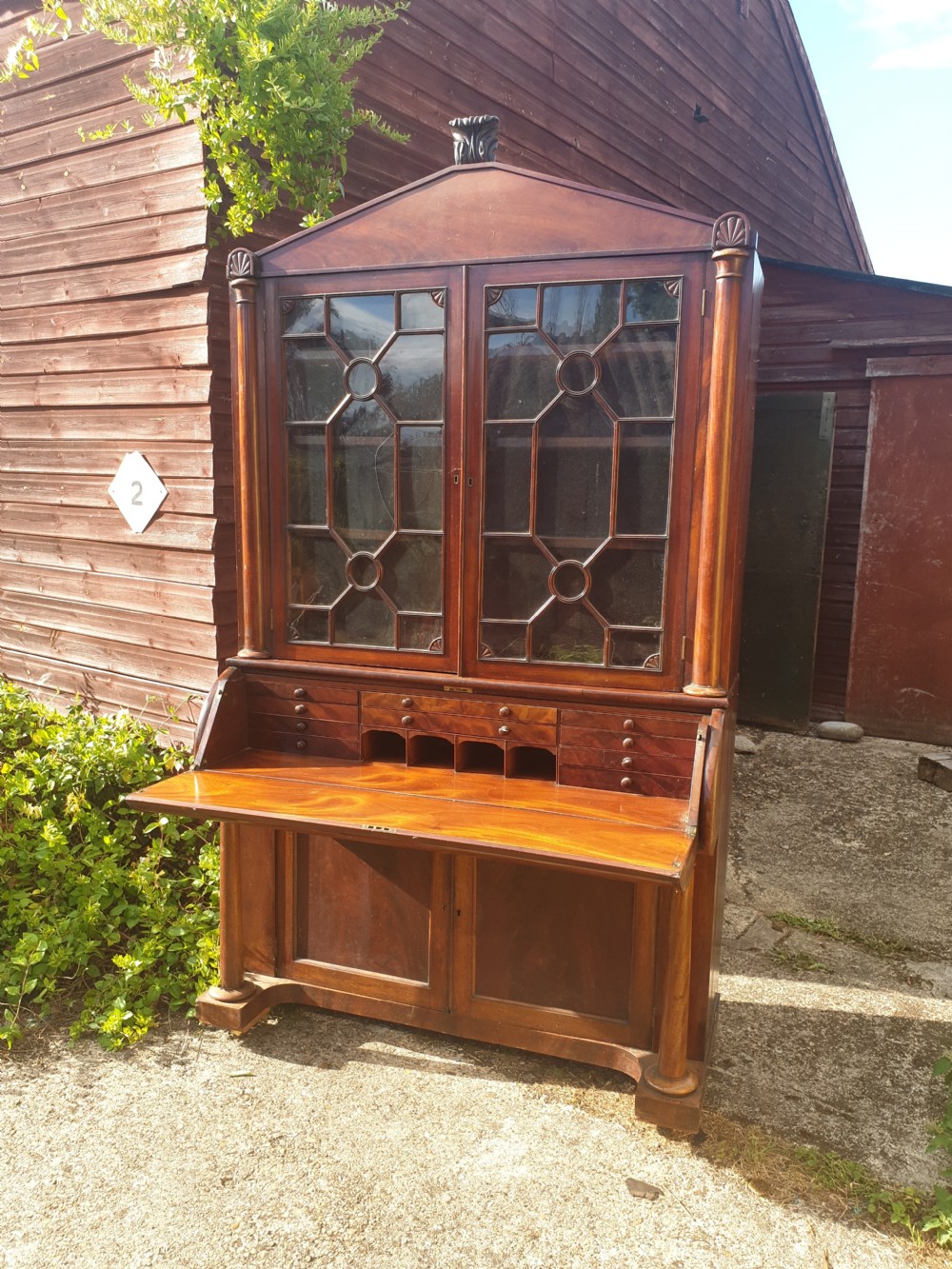 A Regency mahogany bureau bookcase featuring a pediment roof, two glazed cabinet doors with geometric astragal bars, a drop-down desk flap revealing interior drawers, and a lower double-door cupboard flanked by turned side columns.