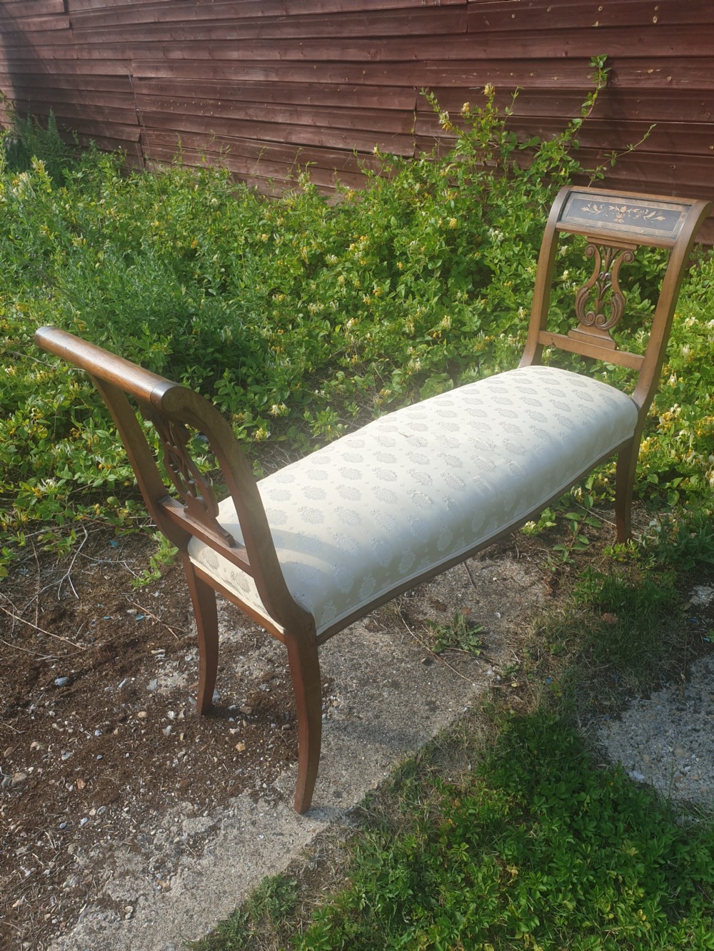 An antique wooden window seat featuring raised ends with lyre-carved splats, floral marquetry top rails, and a cream patterned upholstered cushion. View V