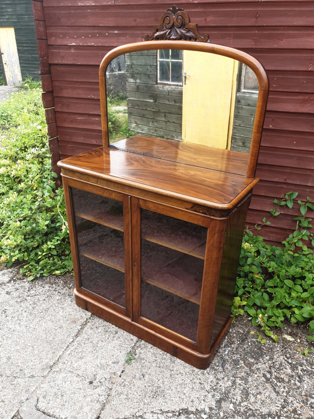 A Victorian warm brown walnut drinks cabinet featuring two glazed doors, internal wooden shelving, and an arched mirror frame topped with a carved cresting.