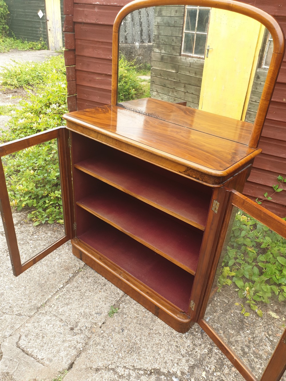 A Victorian warm brown walnut drinks cabinet featuring two glazed doors, internal wooden shelving, and an arched mirror frame topped with a carved cresting. View V