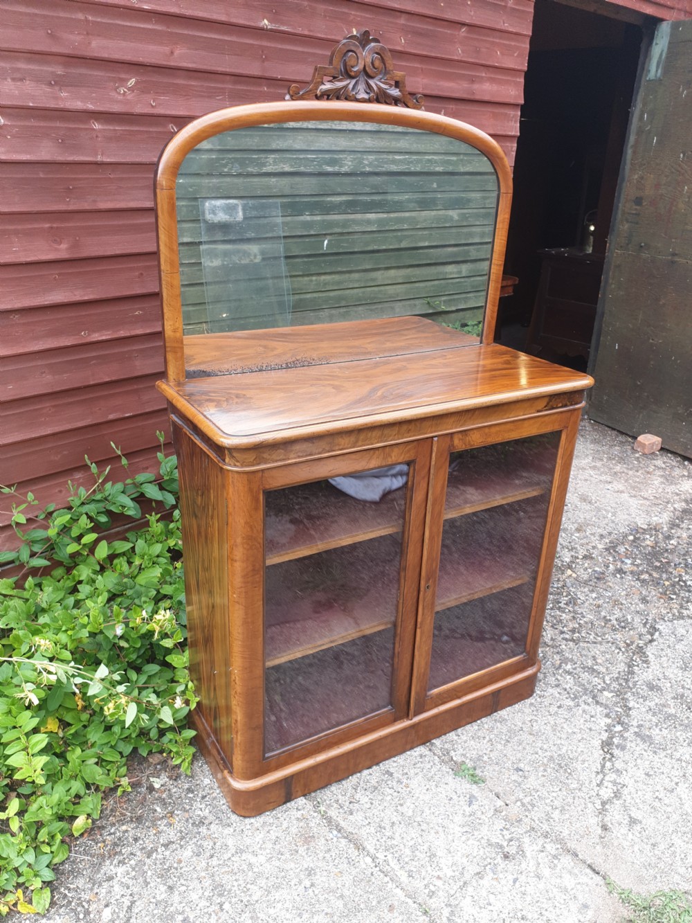 A Victorian warm brown walnut drinks cabinet featuring two glazed doors, internal wooden shelving, and an arched mirror frame topped with a carved cresting. View VI