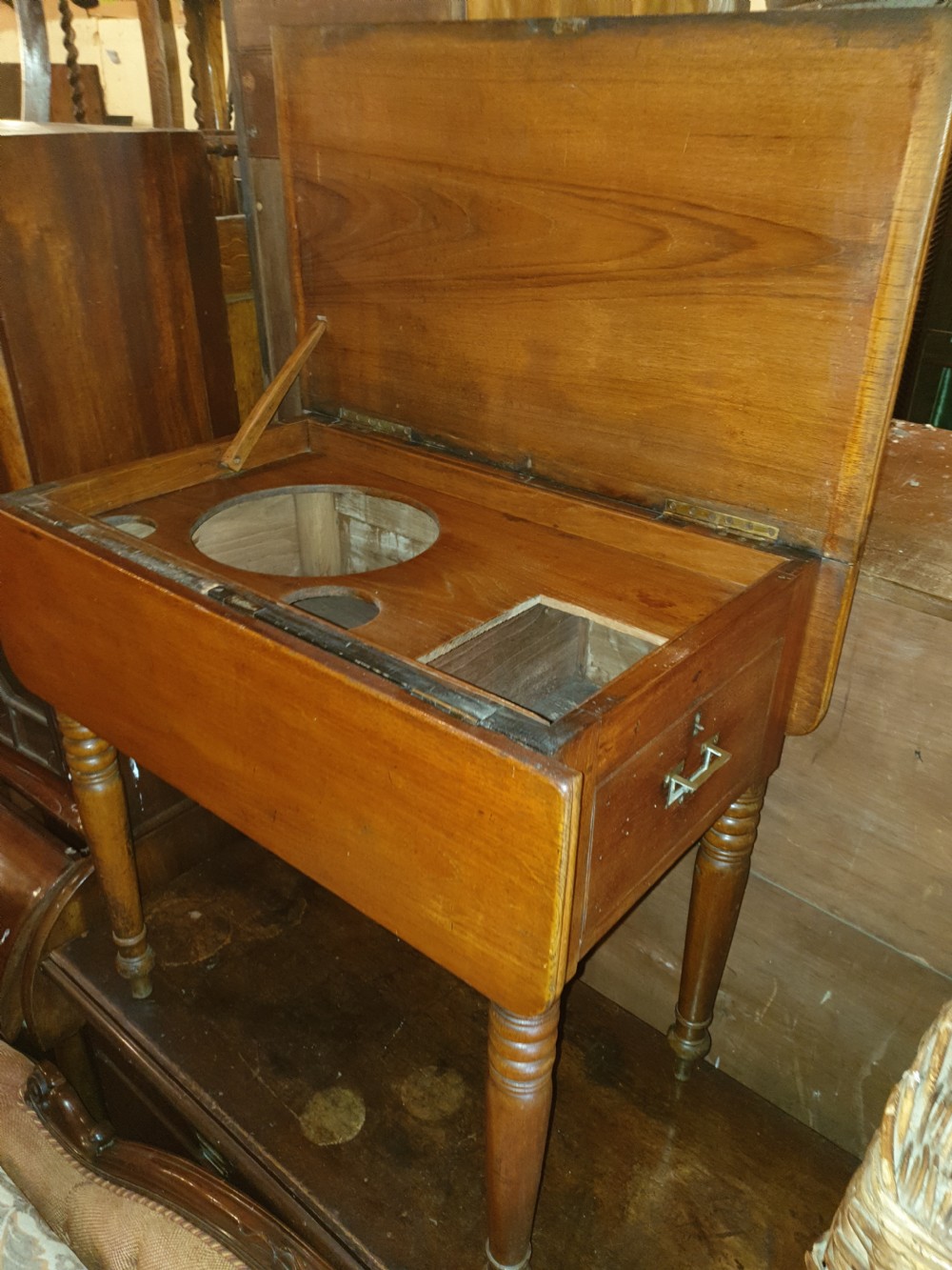 An antique teak ships washstand table featuring a hinged lid opening to reveal circular basin cutouts, resting on turned legs with brass side handles.
