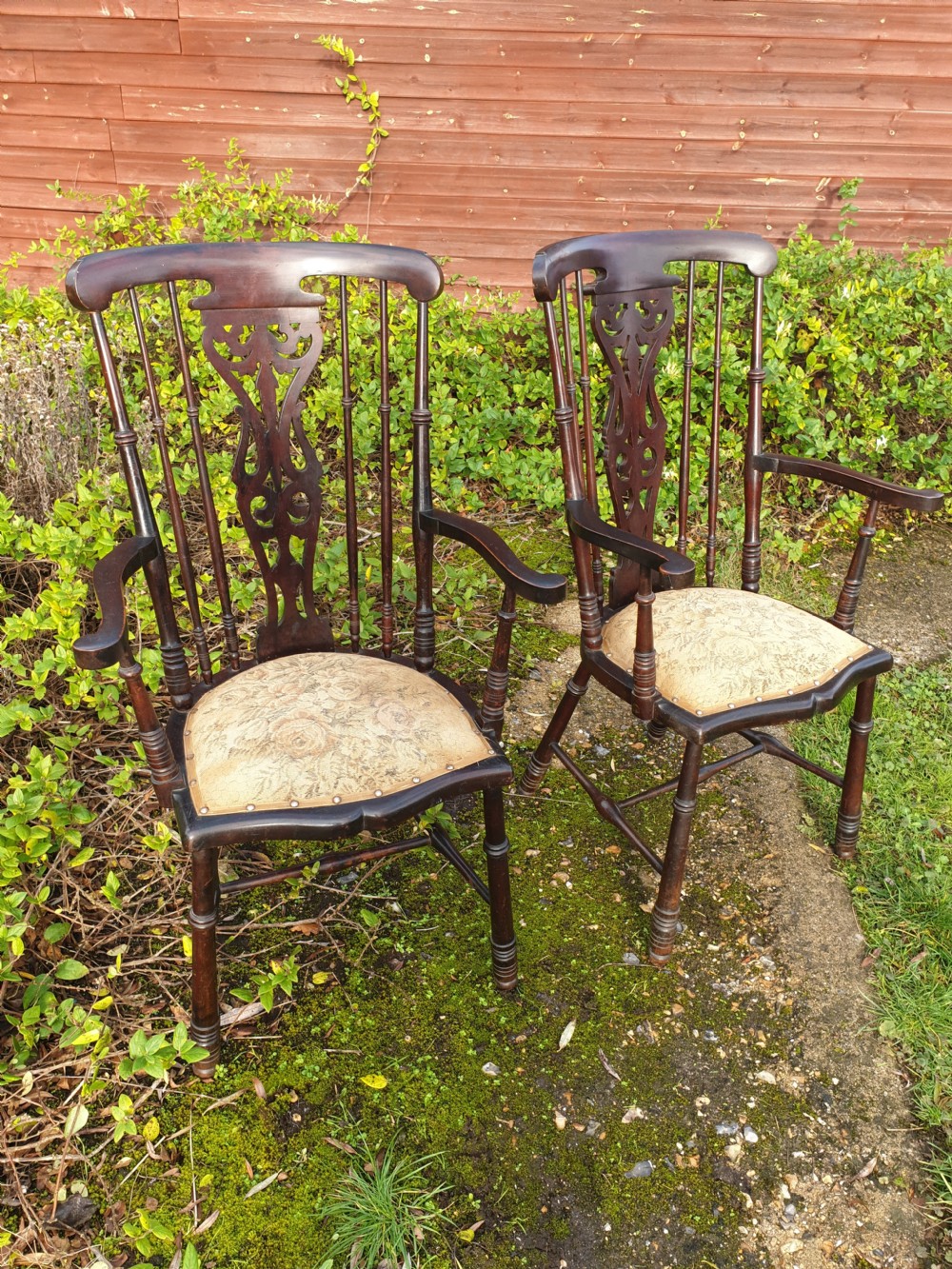 A pair of dark mahogany Edwardian hall armchairs featuring pierced carved back splats, turned spindle supports, curved armrests, and patterned upholstered seats with stud trim.